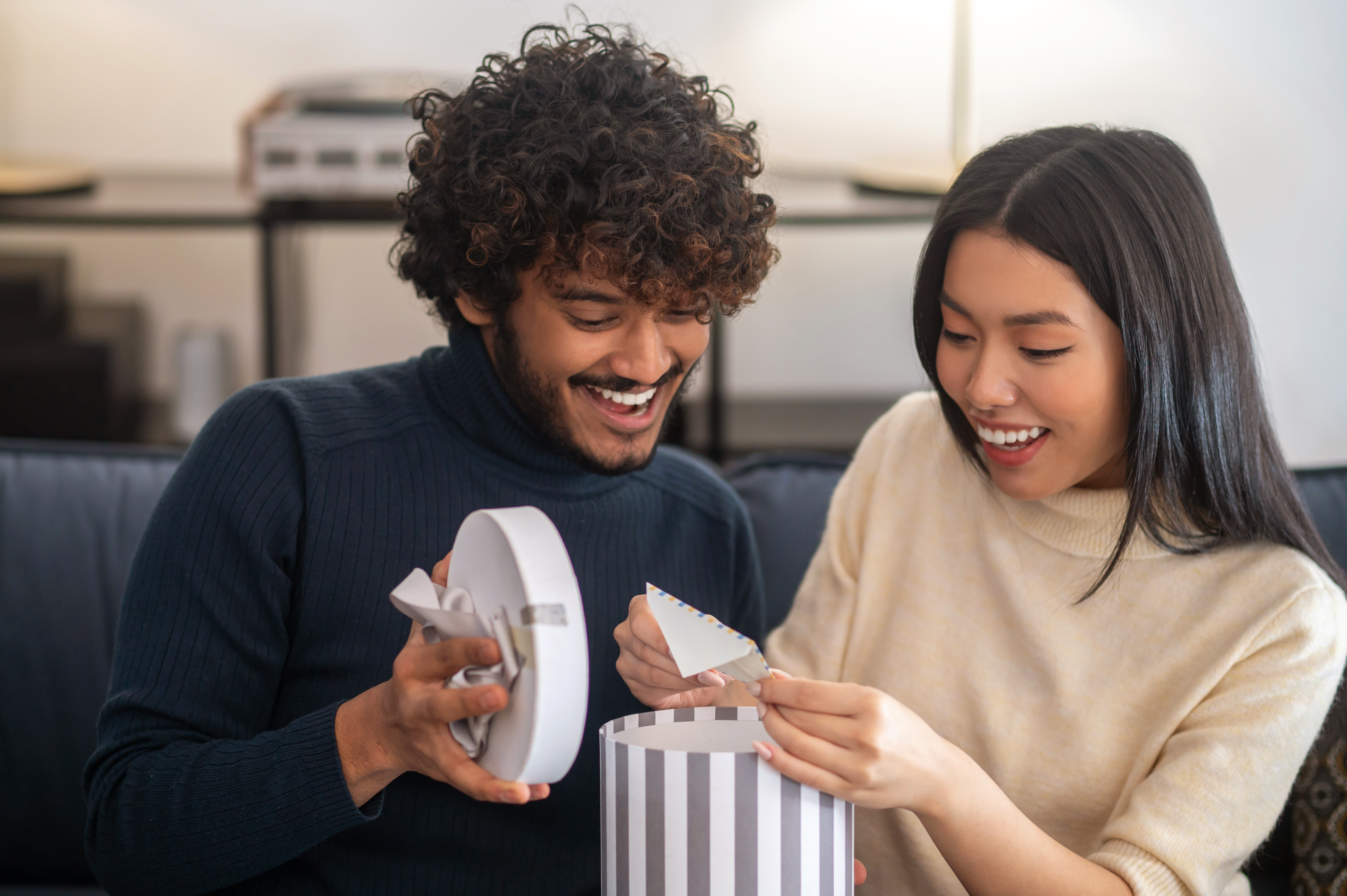 Free photo surprise, joy. smiling attentive curly-haired indian man opening gift light box and pleasantly surprised beautiful asian woman pulling out letter sitting on sofa
